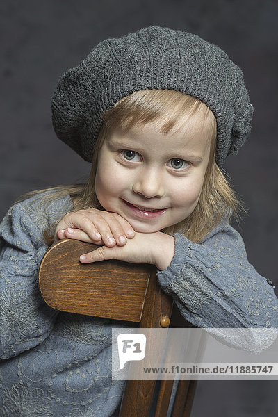 Süßes lächelndes Mädchen mit Strickmütze auf Holzstuhl sitzend vor grauem Hintergrund