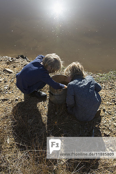 High angle view of boys crouching by bucket at the edge of a river