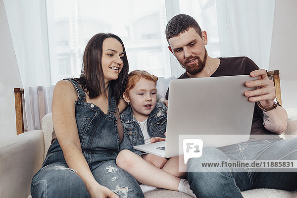 Vater und Mutter mit Tochter schauen auf den Laptop  während sie auf dem Sofa sitzen.