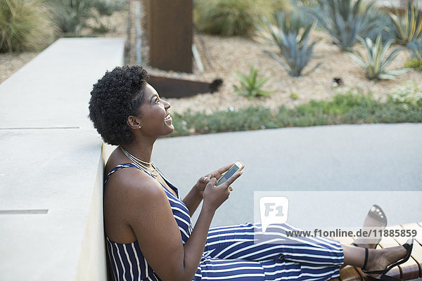 Seitenansicht einer Frau  die sich auf eine Stützmauer stützt  die mit dem Handy sitzt  während sie in den Park blickt.