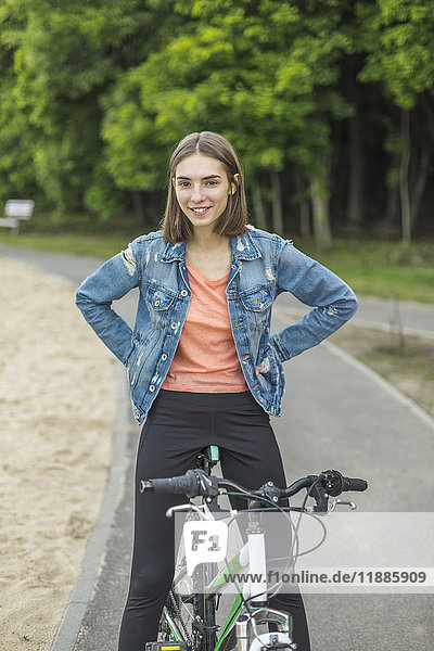 Lächelndes Porträt einer Frau mit Händen an der Hüfte  die auf dem Fahrrad auf der Straße im Park sitzt.
