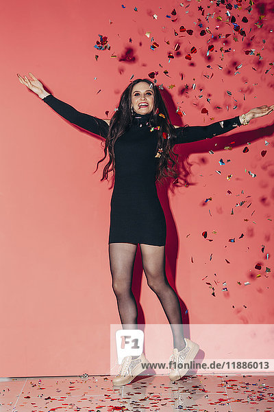 Cheerful young fashionable woman throwing confetti against coral background