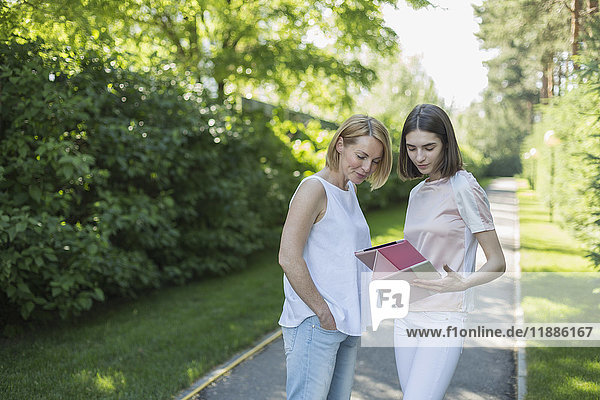 Tochter zeigt der Mutter ein digitales Tablett,  während sie auf einem Fußweg im Park steht.