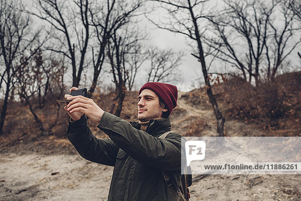 Man with backpack photographing through smart phone while standing against bare trees