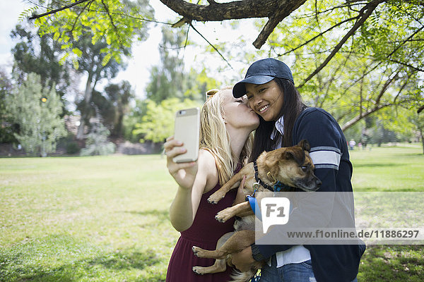 Frau  die sich küsst  während sie Selfie durchs Handy mit Hund im Park nimmt.