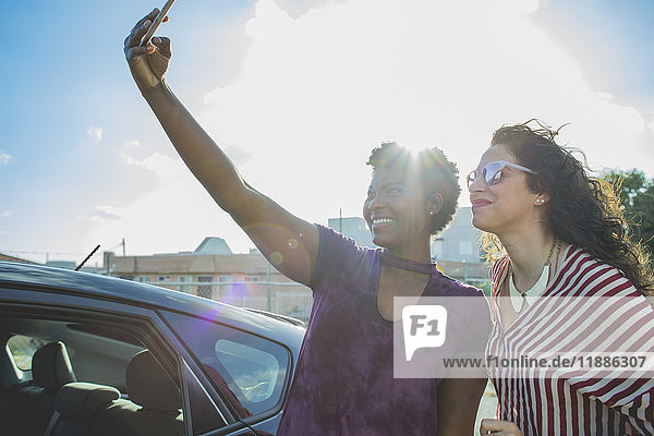 Hinterleuchtete  fröhliche junge Frau nimmt Selfie mit Freundin mit dem Auto gegen den Himmel.