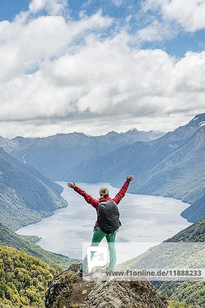 Female hiker is looking at the South Fiord of Lake Te Anau  stretching arms in the air  at back the Southern Alps  the Kepler Track hiking trail  Fiordland National Park  Southland  New Zealand  Oceania