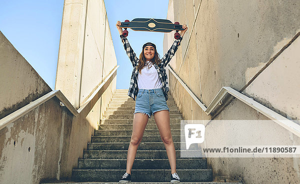 Porträt einer glücklichen jungen Frau  die auf einer Treppe steht und ihr Longboard hochhält.