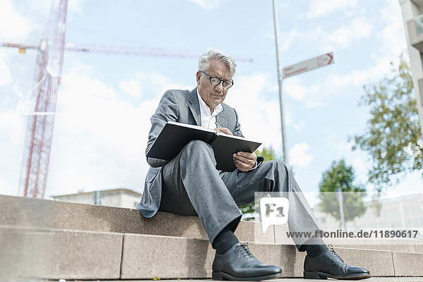 Portrait of senior businessman sitting on stairs with notebook in front of construction crane