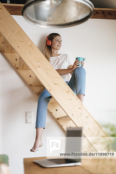 Junge Frau sitzt auf einer Holztreppe  hört Musik und trinkt Tee.