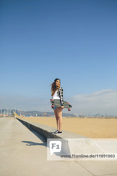 Junge Frau mit Longboard an einer Wand an der Strandpromenade stehend
