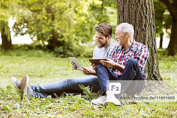 Senior Vater sitzt neben seinem erwachsenen Sohn in einem Park mit Tablette