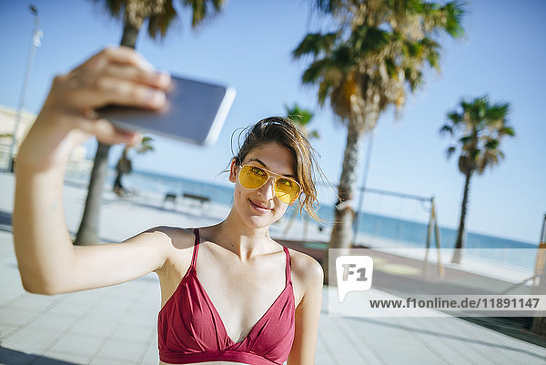 Junge Frau mit gelber Sonnenbrille  die einen Selfie auf der Strandpromenade trägt.