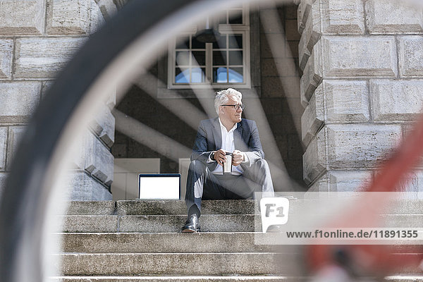 Senior businessman with bicycle sitting on stairs outdoors with laptop and travel mug