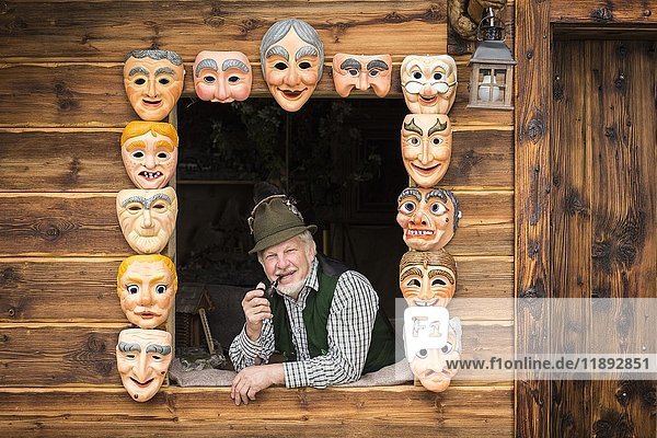 Holzmaskenschnitzer in einem Fenster  eingerahmt von bemalten Holzmasken  Holzmaskenschnitzerei  Bad Aussee  Steiermark  Österreich  Europa