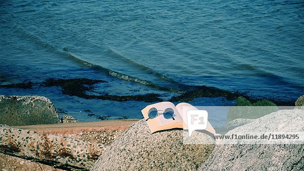 Aufgeschlagenes Buch und Sonnenbrille auf großem Felsen mit sanften Meereswellen  die im Hintergrund an den Strand plätschern