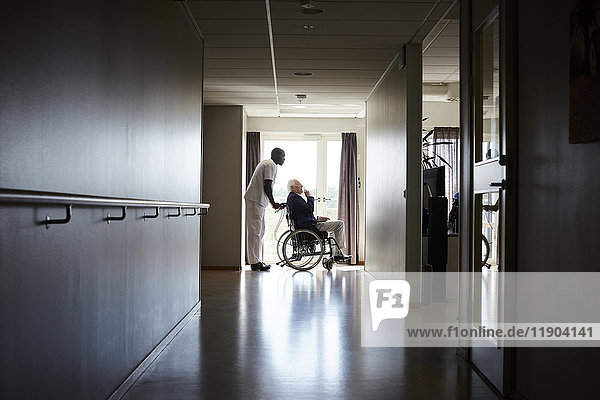 Durchgehende Seitenansicht des Krankenpflegers  der den älteren Mann im Rollstuhl auf dem Flur des Krankenhauses schiebt.