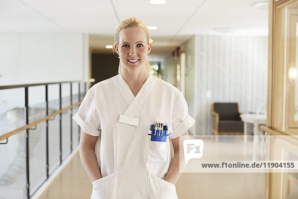 Portrait of happy female nurse standing with hands in pockets at hospital corridor