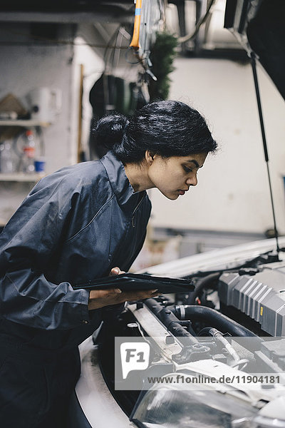 Side view of female mechanic holding digital tablet while examining car engine in auto repair shop