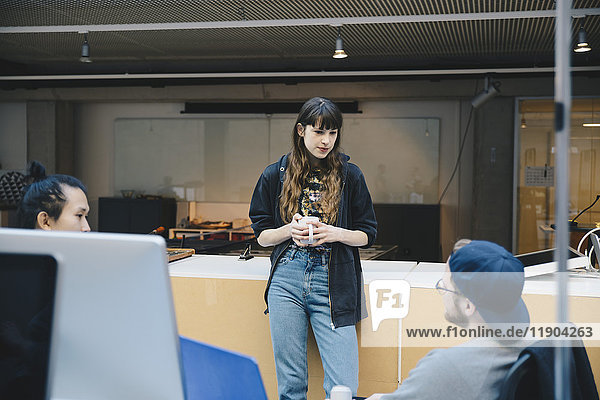 Female computer programmer holding coffee mug while discussing with colleagues in office