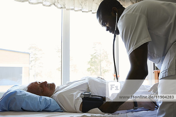 Seitenansicht des Krankenpflegers  der den Blutdruck des älteren Mannes auf dem Krankenhausbett gegen das Fenster prüft.