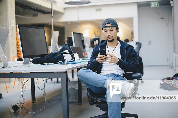 Männlicher Programmierer  der ein Smartphone benutzt  während er im Büro auf einem Stuhl sitzt.