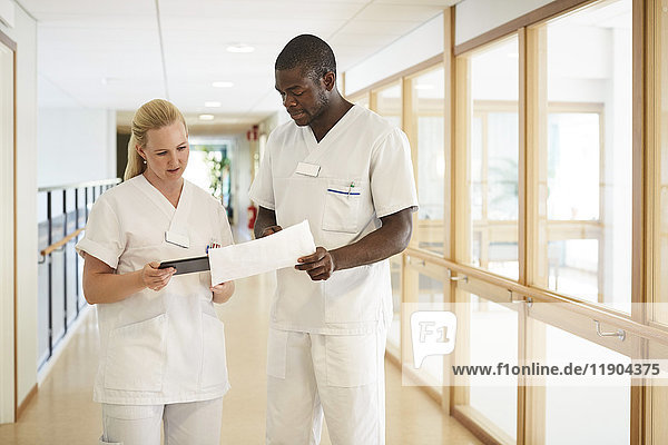 Male and female nurse using digital tablet while examining document in hospital corridor
