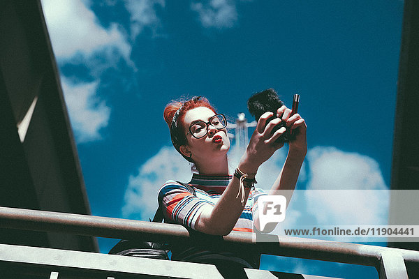 Niederwinkelaufnahme einer jungen Frau,  die sich mit dem Handy an einem Geländer gegen den Himmel lehnt.