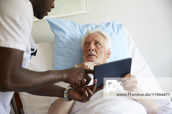 Älterer Mann beim Betrachten des Krankenpflegers bei der Verwendung des digitalen Tabletts auf dem Bett in der Krankenstation.