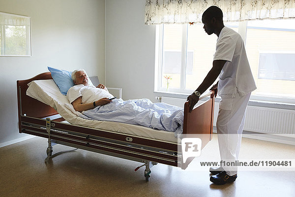 Male nurse adjusting bed while senior man watching TV in hospital ward