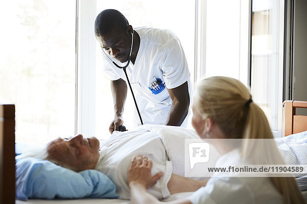 Krankenschwester tröstet älteren Mann  während männlicher Kollege mit Stethoskop auf der Krankenstation untersucht.