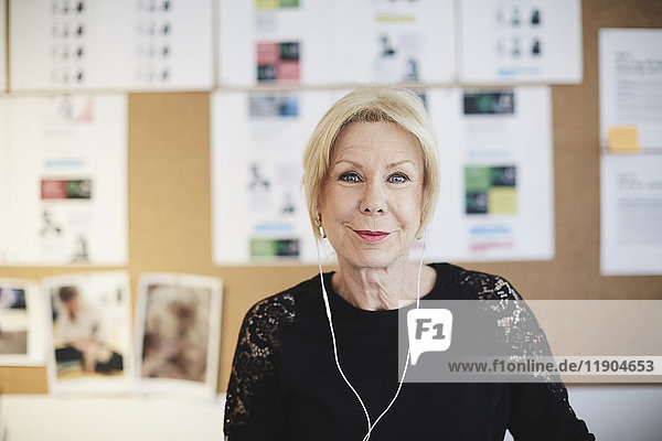 Portrait of smiling senior businesswoman with headphones against bulletin board at creative office