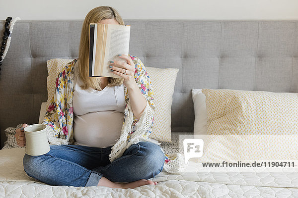 Kaukasische werdende Mutter sitzt auf dem Bett  liest ein Buch und trinkt Kaffee
