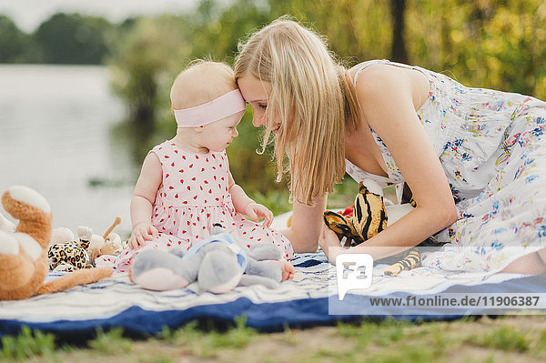 Mutter und Tochter spielen mit Plüschtieren auf einer Decke in der Nähe eines Sees