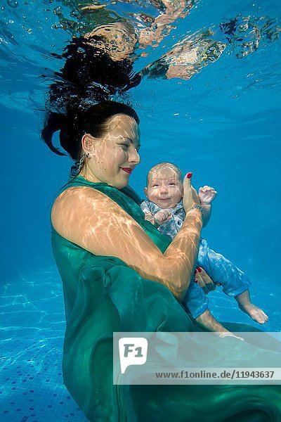 Woman in green dress with little boy posing under water in pool.