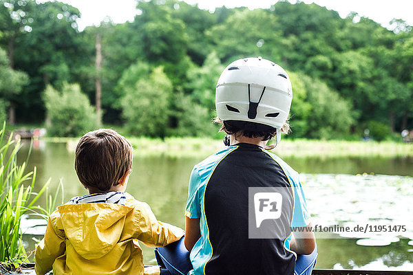 Two young brothers sitting at water's edge  older brother wearing cycling helmet  rear view
