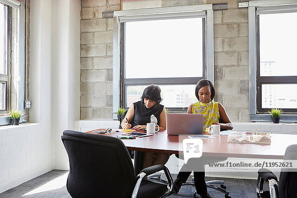 Zwei Frauen sitzen am Tisch des Besprechungszimmers  benutzen einen Laptop und schreiben mit einem Notizbuch