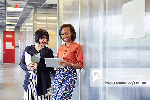 Zwei Geschäftsfrauen stehen im Bürokorridor,  schauen auf das digitale Tablet und lächeln