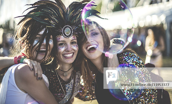 Three young women at a summer music festival wearing feather headdress and faces painted  smiling at camera.