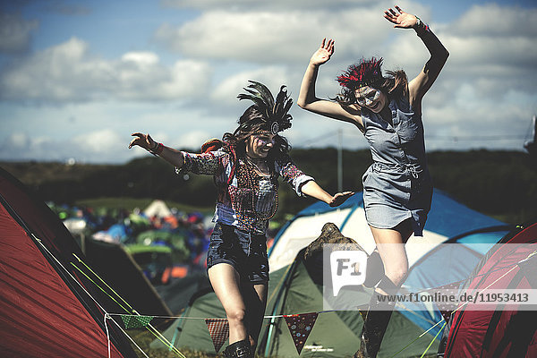 Zwei lächelnde junge Frauen bei einem Sommer-Musikfestival mit geschminktem Gesicht,  Federkopfschmuck tragend,  zwischen Zelten hüpfend.