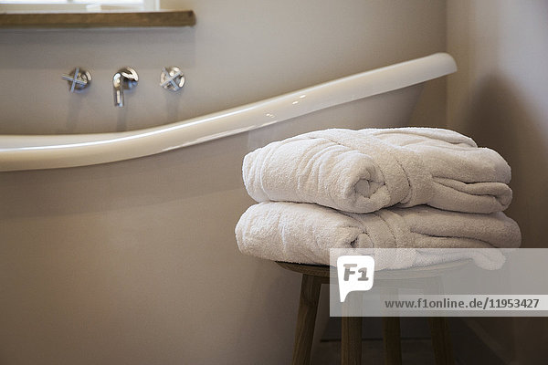 Eine altmodische Badewanne in Pantoffelform  Badewanne mit erhöhtem Ende und wandmontierten Wasserhähnen in einem Badezimmer. Zwei gefaltete Gästebademäntel auf einem Hocker.