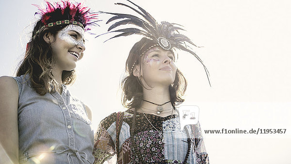 Zwei junge Frauen bei einem Sommer-Musikfestival mit bemalten Gesichtern und Federkopfschmuck.