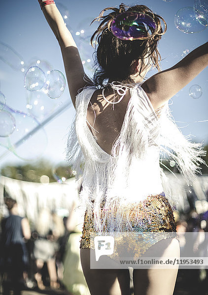 Rückansicht einer jungen Frau bei einem Sommer-Musikfestival,  die in goldenen,  mit Pailletten bestickten Hotpants unter der Menge tanzt.