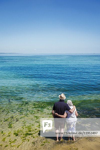 Rückansicht eines älteren Ehepaars  das mit Armen um die Schultern steht und über das lebendige  klare  blaue Wasser auf das Meer blickt.