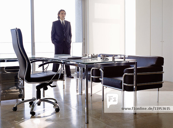 Geschäftsmann in dunklem Anzug  der drinnen in einem Büro hinter einem Schreibtisch mit Stühlen steht.