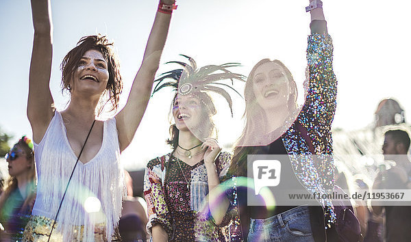Three smiling young women at a summer music festival face painted  wearing feather headdress  arms raised  standing among the crowd.
