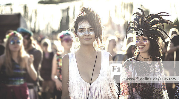 Zwei lächelnde junge Frauen bei einem Sommer-Musikfestival mit bemaltem Gesicht  mit Federkopfschmuck  stehen in der Menge und schauen in die Kamera.
