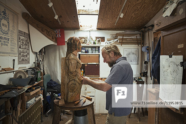 Ein Holzschnitzer  der in seiner Werkstatt steht und mit Handwerkzeugen eine hölzerne weibliche Schiffsgestalt aus Holz formt und verziert.