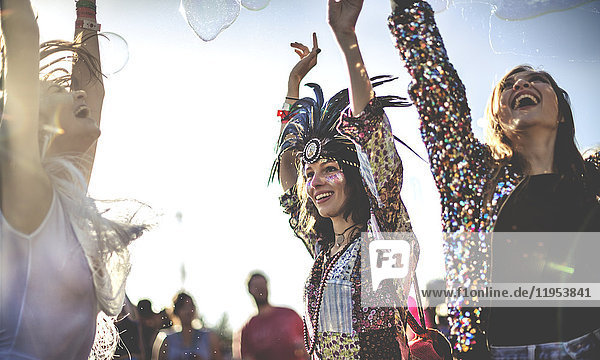 Drei junge Frauen bei einem Sommer-Musikfestival mit Federkopfschmuck und Gesichtsbemalung  erhobenen Armen und tanzend.