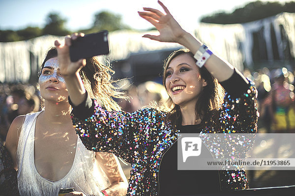 Junge lächelnde Frau bei einem Sommer-Musikfestival  trägt eine mehrfarbige Paillettenjacke und fotografiert mit einem Smartphone.
